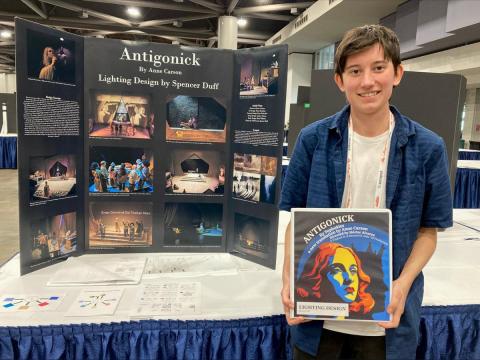 Spencer Duff poses for a photo holding with a binder and standing in front of a poster board of the play Antigonick