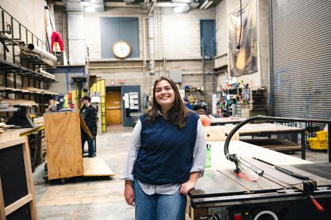 Avery McGarry stands in the scene shop wearing a blue sweater vest and smiling at the camera.